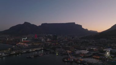Deniz kıyısındaki yat limanının üzerinden uç. Akşam kasabasının binaları ve caddeleri ve alacakaranlıkta gökyüzüne karşı dağların silueti. Cape Town, Güney Afrika.