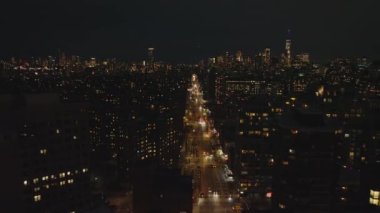 Aydınlanmış caddenin üzerinde ilerleyip yerleşim yerlerinin olduğu mahalleye doğru ilerliyorlar. Arka planda yüksek katlı iş binaları olan bir gece şehri sahnesi. Manhattan, New York City, ABD.