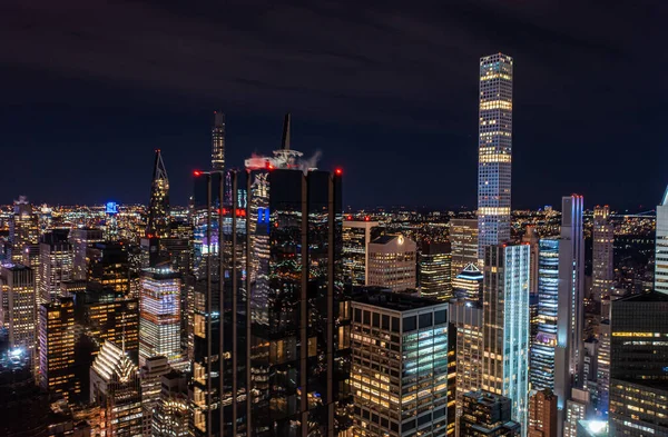 Night city scene with modern high rise buildings in metropolis. Dark glossy glass facade reflecting surrounding lights. Manhattan, New York City, USA.