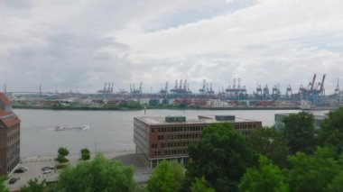 Buildings on wide river waterfront. Forwards fly above vegetation, revealing passenger ship passing on Elbe river along large cargo harbour. Hamburg, Germany.