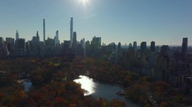 Sonbahar Central Park 'ın üzerinde renkli ağaçlar ve güneşi yansıtan su yüzeyiyle uçun. Arka planda modern şehir merkezi ofis kuleleri var. Manhattan, New York City, ABD.
