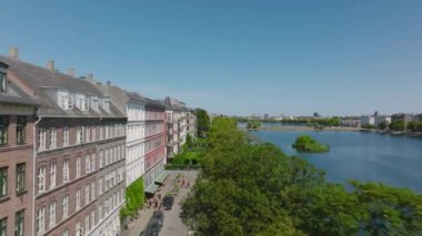 Göl kıyısındaki yemyeşil ağaçların üzerinde ilerliyorlar. Şehrin rıhtımındaki evlerde sıra sıra. Açık mavi gökyüzü ile güneşli bir gün. Kopenhag, Danimarka.