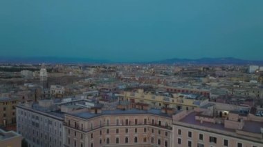 Akşam kentinde binaların üzerinde ileriye doğru uçar. Alacakaranlıkta şehir merkezinde çatı terasları olan çok katlı apartmanlar. Roma, İtalya.