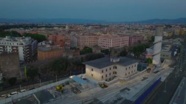 Demiryollarını ve apartman bloklarını ayıran antik şehir duvarlarının üzerinden uçmak. Alacakaranlıkta çatı teraslarının üstünde alçak uçuş. Roma, İtalya.