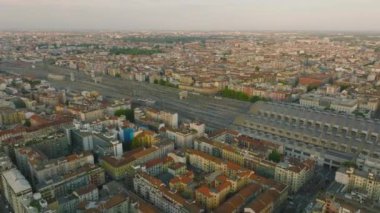 Gün batımında büyük şehrin havadan panoramik görüntüsü. Tren istasyondan kalkıyor. Konut binalarıyla çevrili geniş demiryolu koridoru. Milano, İtalya