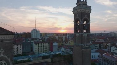 Şehir merkezindeki binaların renkli günbatımı gökyüzüne karşı hava görüntüleri. Tersten bakınca su kanalı üzerinde iki kulesi olan tuğladan bir kilise görülüyor. Milano, İtalya.
