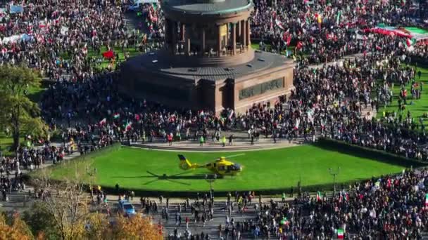 イランの抗議デモ中に広場に人々の群衆の空中ビュー 緑の芝生の上に立って黄色の救助ヘリコプター ドイツ ベルリン — ストック動画