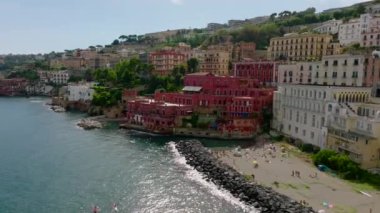 Turizm merkezindeki deniz kıyısındaki güzel lüks sarayların hava kaydırak ve pan görüntüleri. İnsanlar kumsalda güneşli bir günün tadını çıkarıyor. Tarihi binaların renkli cepheleri. Napoli, İtalya.