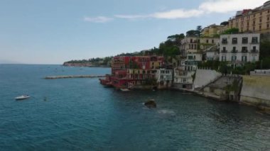 Deniz kıyısındaki renkli binaların etrafında ileriye doğru uçar, insanlar kıyıda suda yüzer. Napoli, İtalya.