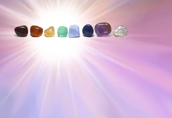 Chakra coloured tumbled healing crystal healing stones template -  bright white light burst on a pink purple graduated background with a row of crystals top left and copy space beneath