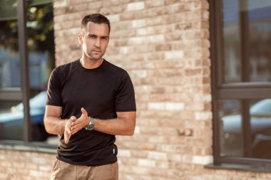 Siyah tişörtlü, kısa tüylü yakışıklı adam tuğla bir duvarın arkasında.