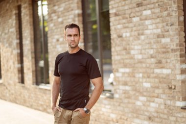 Siyah tişörtlü, kısa tüylü yakışıklı adam tuğla bir duvarın arkasında. 