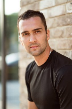 Siyah tişörtlü, kısa tüylü yakışıklı adam tuğla bir duvarın arkasında.