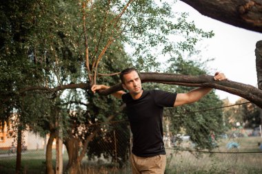 Elinde ağaç dalı tutan, siyah tişörtlü, hafif tüylü bir gencin portresi.