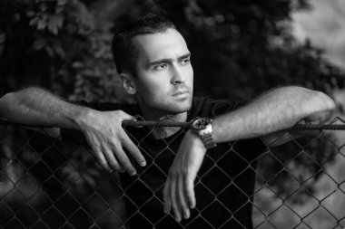 Tıraşsız, siyah tişörtlü, tel örgüye yaslanmış yakışıklı bir adamın portresi.