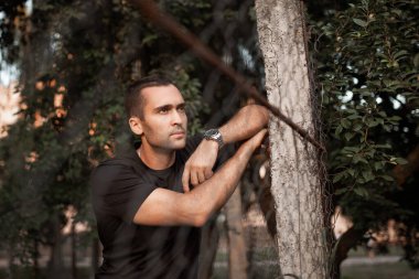 Tıraşsız, siyah tişörtlü, tel örgüye yaslanmış yakışıklı bir adamın portresi.