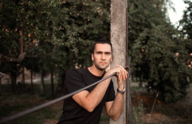 Tıraşsız, siyah tişörtlü, tel örgüye yaslanmış yakışıklı bir adamın portresi.