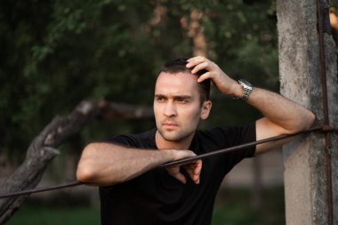 Tıraşsız, siyah tişörtlü, tel örgüye yaslanmış yakışıklı bir adamın portresi.