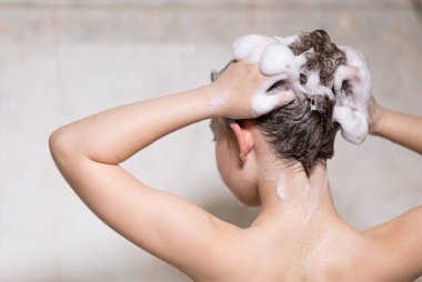 Küçük, güzel bir kız, beyaz bir banyoda sabun köpükleriyle oturuyor, şampuan yapıyor ve başını saçlarıyla yıkıyor. Model fotoğrafçılık.