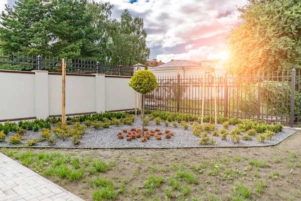 Arka ve ön bahçe kır evi bahçesi, çiçek açan bitki ve yeşil çimenler, kahverengi kaldırım ve turuncu tuğla duvar, arka planda her zaman yeşil ağaçlar, parktaki bakımlı peyzaj.