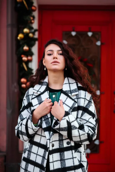 Stylish woman in checkered coat in front of the red door with Christmas festive decorations — Stock Photo, Image