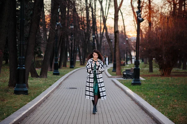 Fashionable woman in checkered elegant coat and green dress walking in park in autumn or in winter. — Stock Photo, Image