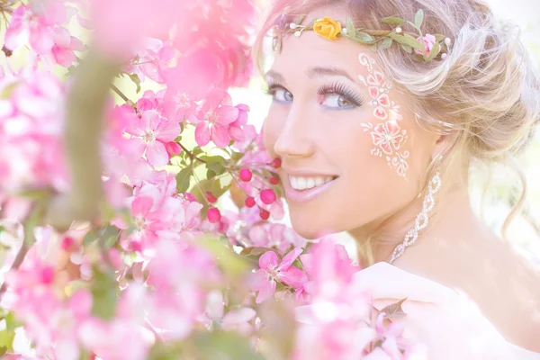 Unga vackra eleganta och attraktiva flicka står i en skog nära blommande träd med långa hår blond i solig dag och smink bland blommor med blå ögon — Stockfoto