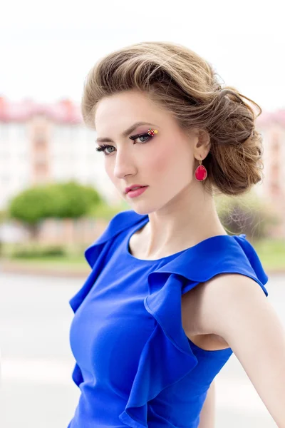 Beautiful young girl in a blue dress with a beautiful hairstyle and makeup stands on street in town — Stock Photo, Image