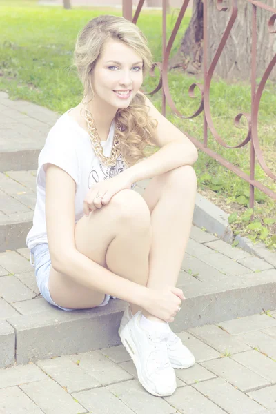 Bella ragazza con un sorriso, seduta sulle scale in pantaloncini, scarpe da ginnastica in un parco in una luminosa giornata soleggiata — Foto Stock