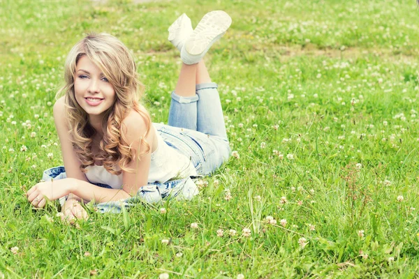 Feliz joven hermosa chica sexy tumbada en la hierba y sonríe en jeans en un día soleado de verano en el jardín — Foto de Stock