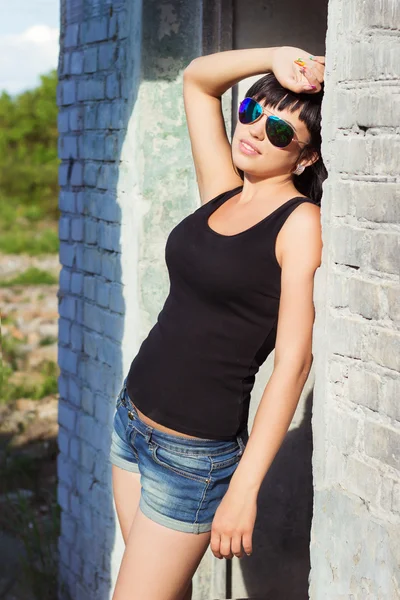 Hermosa chica sexy joven con el pelo largo en un día soleado de verano en pantalones cortos de pie al aire libre en gafas de sol cerca de paredes de ladrillo blanco — Foto de Stock