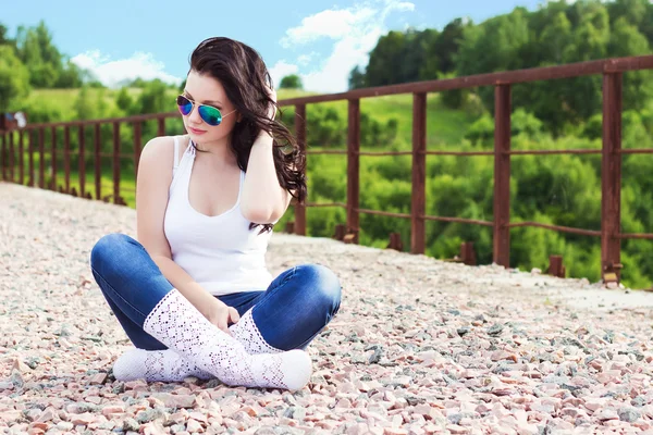 Mooi meisje met lange zwarte haren in een zonnige zomerdag zittend op de brug in zonnebril — Stockfoto