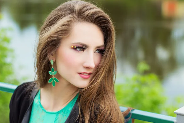 Joven hermosa chica sexy en un vestido verde con hermoso maquillaje con grilletes verdes sentado en la orilla del río en la ciudad — Foto de Stock