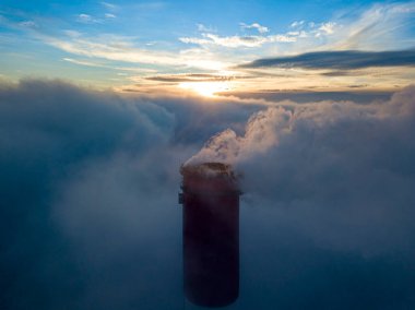 Gün batımında sislerin üzerinde bir termik santralin yüksek bacası. Hava aracı görünümü.