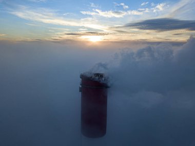 Gün batımında sislerin üzerinde bir termik santralin yüksek bacası. Hava aracı görünümü.