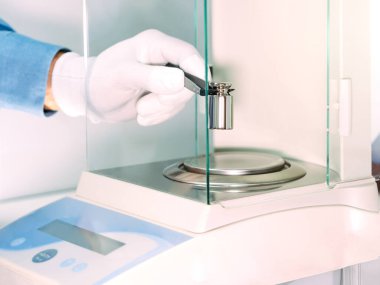 Bir operatörün eli analitik dengeyi sağlamak için çelik kalibrasyon ağırlığını tutuyor. Laboratuvarda kalite kontrolü kavramı.