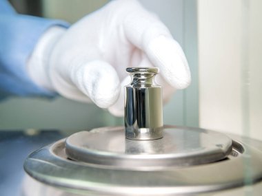 Bir operatörün eli analitik dengeyi sağlamak için çelik kalibrasyon ağırlığını tutuyor. Laboratuvarda kalite kontrolü kavramı.