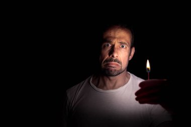 Karanlıkta elinde kibritle kameraya bakan endişeli bir adam. Karartma konsepti.