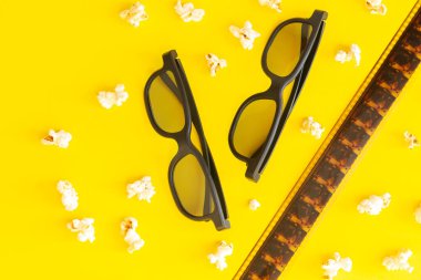 Film zamanı konsepti, patlamış mısırlı sinema arka planı, üç boyutlu gözlükler ve sarı masanın üstündeki film şeridi üst görüntüsü.