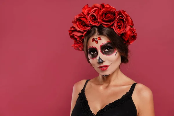 Krásná dívka v la muerta oblečení a lebka tvoří při pohledu do kamery během halloween izolované na růžovém pozadí. — Stock fotografie