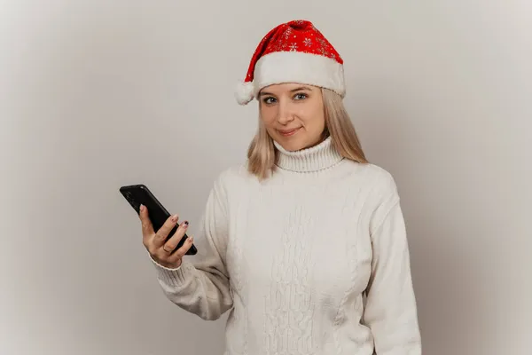 Woman in a white sweater and santa hat with a phone in her hands on a gray isolated background — Stock Photo, Image