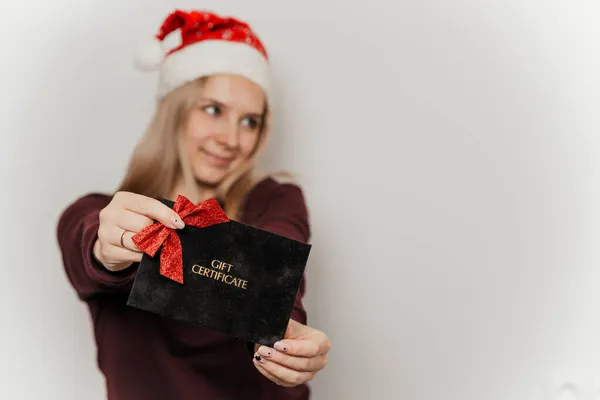 Woman in a burgundy sweater and red santa hat holding black gift certificate on a gray background — Stock Photo, Image