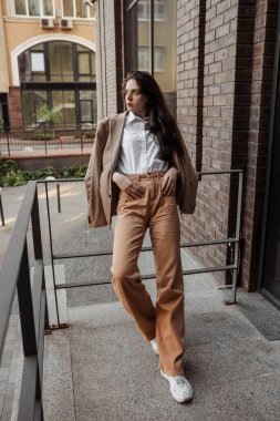  Korkulukların yanında poz veren, büyük ceketli, uzun boylu, şık bir kız ve şehrin arka planına basıyor.