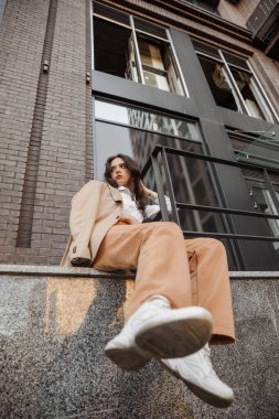  Korkulukların yanında poz veren, büyük ceketli, uzun boylu, şık bir kız ve şehrin arka planına basıyor.