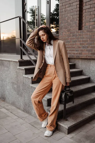 Chica alta y elegante con una bolsa en una chaqueta beige de gran tamaño y pantalones naranja en el fondo de la pared de ladrillo — Foto de Stock