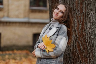  Gri ceketli, gülümseyen genç kız elinde sarı bir yaprak tutuyor.