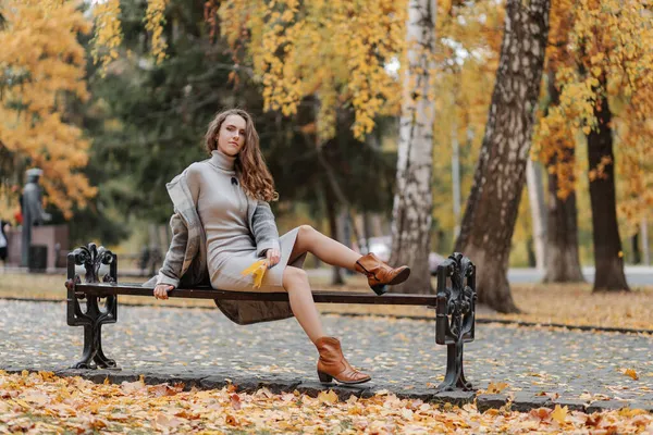 Joven chica de pelo rizado en un vestido gris se sienta en un banco en un parque de otoño —  Fotos de Stock