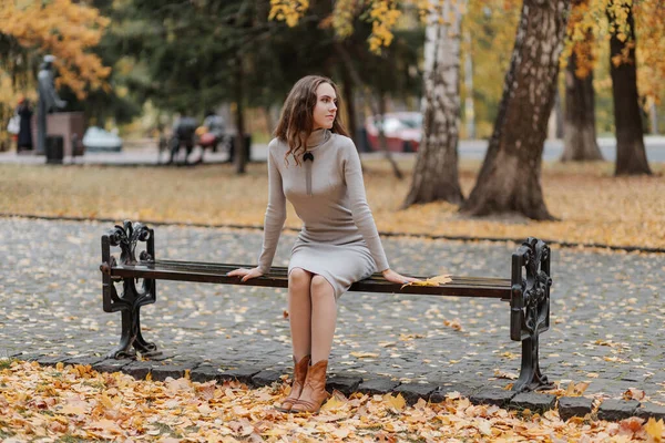 Giovane ragazza dai capelli ricci in un abito grigio si siede su una panchina in un parco autunnale — Foto Stock