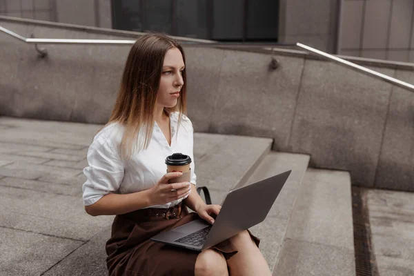 Молода ділова жінка тримає каву та ноутбук на фоні сучасного офісного будівництва — стокове фото