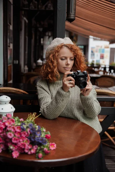 Una donna con un berretto e un maglione verde tiene una macchina fotografica tra le mani e scatta — Foto Stock